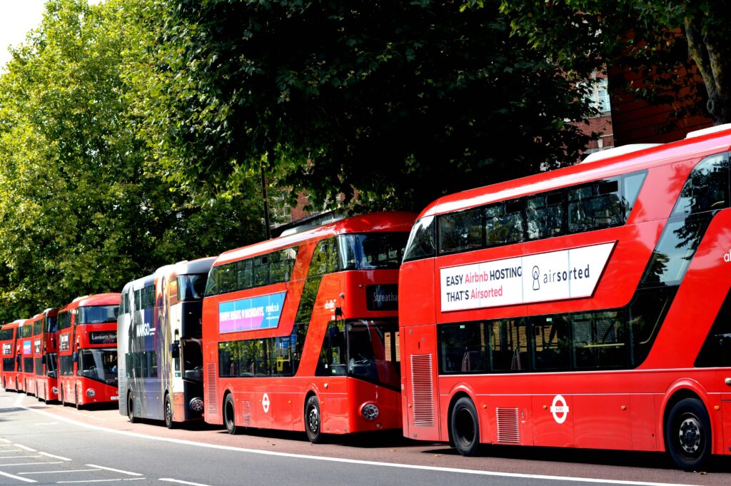 A row of traditional red double-decker buses parked on a London street under green trees.